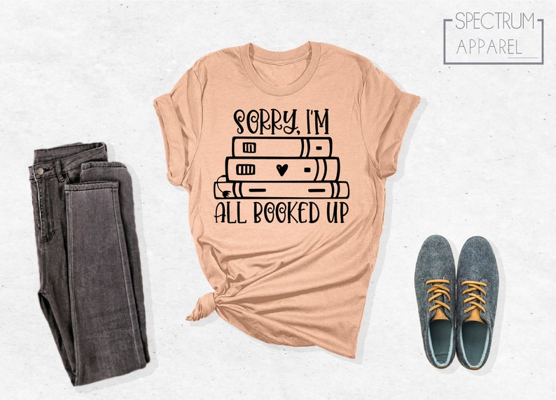 May include: A peach-colored t-shirt with a black graphic of a stack of books and the text "Sorry, I'm all booked up".
