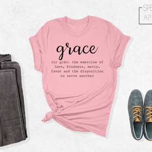 Op de afbeelding: Een roze T-shirt met het woord "grace" in cursief en een definitie van het woord eronder. De definitie luidt: "(n) gr&auml;s: de uitoefening van liefde, vriendelijkheid, genade, gunst en de bereidheid om een ander te dienen".