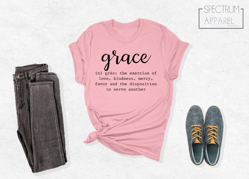 Op de afbeelding: Een roze T-shirt met het woord "grace" in cursief en een definitie van het woord eronder. De definitie luidt: "(n) gr&auml;s: de uitoefening van liefde, vriendelijkheid, genade, gunst en de bereidheid om een ander te dienen".