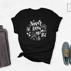 May include: Black t-shirt with white text that says "Never Grow Up" surrounded by white illustrations of Disney park icons.