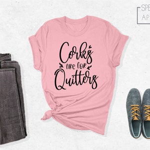 May include: A pink t-shirt with the text "Corks are for Quitters" in black lettering. The shirt is folded and tied in a knot. A pair of dark gray jeans and two gray shoes are also in the image.