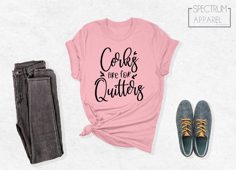 May include: A pink t-shirt with the text "Corks are for Quitters" in black lettering. The shirt is folded and tied in a knot. A pair of dark gray jeans and two gray shoes are also in the image.