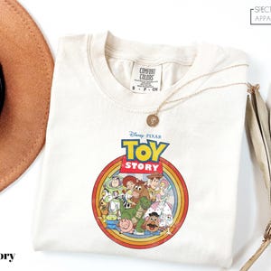a white t-shirt with a Disney Pixar logo, specifically the characters from the Toy Story movie, displayed on it. The t-shirt is laid out on a white surface, with a brown hat and a fan visible in the background.