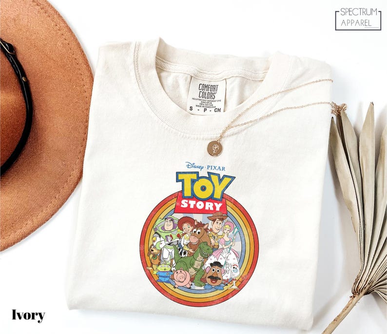 a white t-shirt with a Disney Pixar logo, specifically the characters from the Toy Story movie, displayed on it. The t-shirt is laid out on a white surface, with a brown hat and a fan visible in the background.