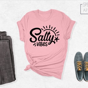 May include: A pink t-shirt with the text "Salty Vibes" in black lettering and a sun graphic. The shirt is laid flat on a white background with a pair of dark grey jeans and a pair of grey shoes with orange laces.