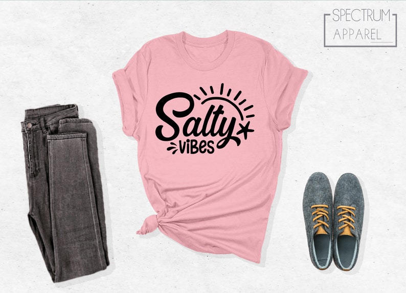 May include: A pink t-shirt with the text "Salty Vibes" in black lettering and a sun graphic. The shirt is laid flat on a white background with a pair of dark grey jeans and a pair of grey shoes with orange laces.