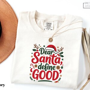 May include: Ivory-colored Comfort Colors t-shirt with the text "Dear Santa, define GOOD" in red and green. The design includes a Santa hat, holly, and snowflakes. A gold necklace is draped across the shirt.
