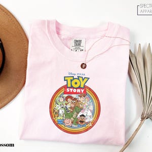 a pink t-shirt with a Disney Pixar logo, specifically the characters from the Toy Story movie, displayed on it. The t-shirt is placed on a white surface, with a brown hat and a plant visible in the background.