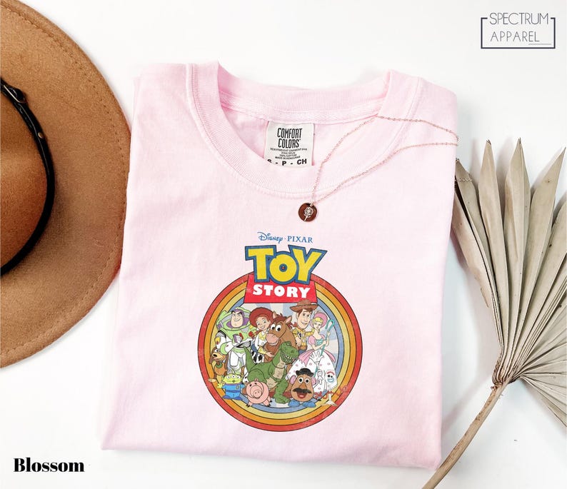 a pink t-shirt with a Disney Pixar logo, specifically the characters from the Toy Story movie, displayed on it. The t-shirt is placed on a white surface, with a brown hat and a plant visible in the background.