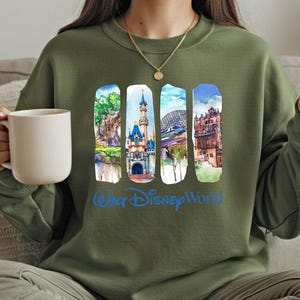 A woman wearing a green sweatshirt with a collage of Disney World images on it, holding a white mug.