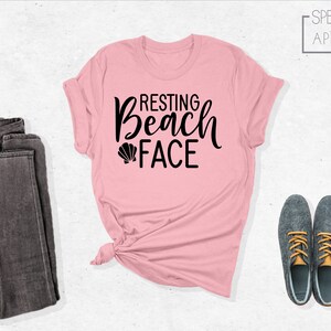 Może przedstawiać: R&oacute;żowa koszulka z czarnym napisem "Resting Beach Face" i małym grafiką muszelki.