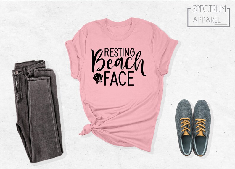Może przedstawiać: R&oacute;żowa koszulka z czarnym napisem "Resting Beach Face" i małym grafiką muszelki.