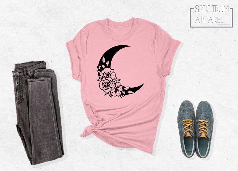 May include: A pink t-shirt with a black crescent moon design with floral accents. The design features a crescent moon with a floral arrangement around the bottom of the moon.