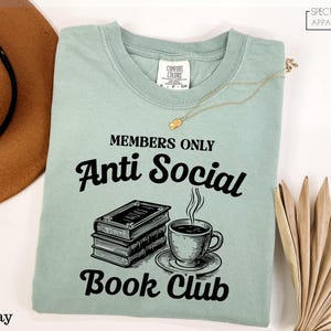 a light green t-shirt with a graphic design and text, along with a hat, a necklace, and some books, all laid out on a white background.