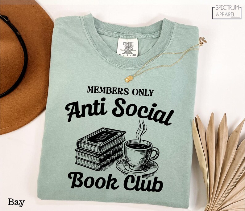 a light green t-shirt with a graphic design and text, along with a hat, a necklace, and some books, all laid out on a white background.