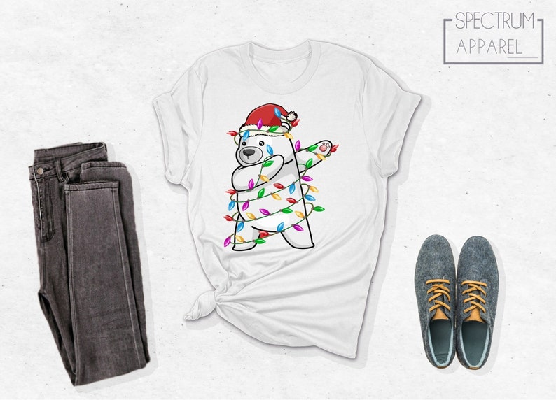 Op de afbeelding: Wit T-shirt met een cartoon ijsbeer met een kerstmuts en gewikkeld in kleurrijke kerstverlichting. De beer doet een dab. Het shirt wordt gecombineerd met een grijze jeans en grijze schoenen.