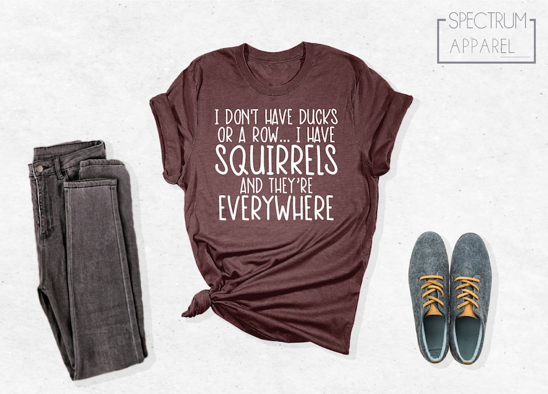 May include: A maroon t-shirt with white text that reads "I don't have ducks or a row... I have squirrels and they're everywhere". The shirt is tied in a knot at the bottom. The text "SPECTRUM APPAREL" is in white on a black background in the upper right corner of the image.