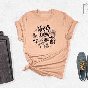 May include: A peach coloured t-shirt with a black graphic that says "Never Grow Up" surrounded by Disney park icons.