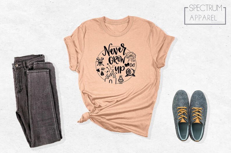 May include: A peach coloured t-shirt with a black graphic that says "Never Grow Up" surrounded by Disney park icons.