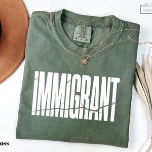 May include: A moss green t-shirt with the word "IMMIGRANT" printed in large white letters. The text has a distressed, cracked effect. A gold necklace with a pendant rests on the shirt. The shirt is labeled "Comfort Colors".