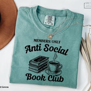 a light green t-shirt with a graphic design that reads "MEMBERS ONLY Anti Social Book Club" and includes a book and a cup of coffee.