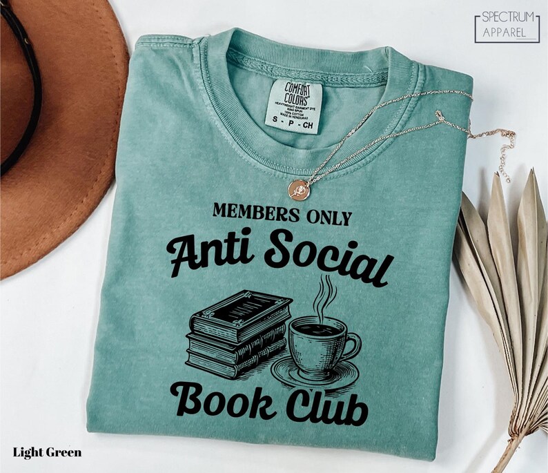 a light green t-shirt with a graphic design that reads "MEMBERS ONLY Anti Social Book Club" and includes a book and a cup of coffee.