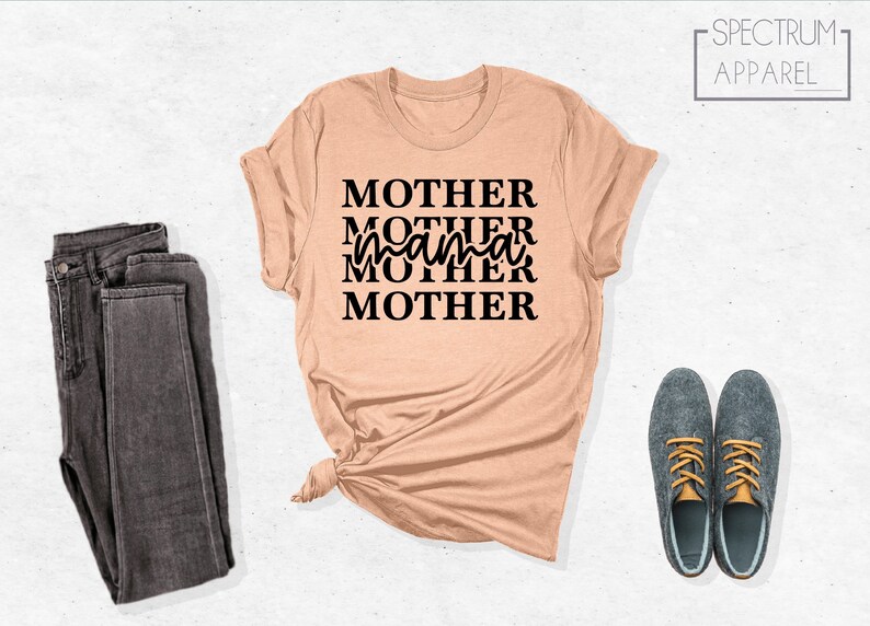 Puede incluir: Una camiseta de color melocot&oacute;n con el texto "MOTHER MOTHER mama MOTHER MOTHER" en negro. La camiseta se muestra con un par de vaqueros gris oscuro y dos zapatos grises con cordones naranjas.
