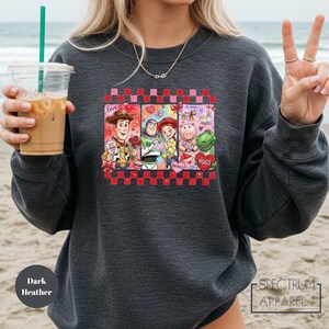 A woman wearing a gray sweatshirt with a graphic of characters from the Toy Story movie, including Woody, Buzz Lightyear, and other beloved characters, along with a peace sign. She is holding a drink in a plastic cup with a straw.