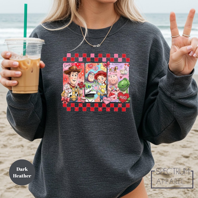 A woman wearing a gray sweatshirt with a graphic of characters from the Toy Story movie, including Woody, Buzz Lightyear, and other beloved characters, along with a peace sign. She is holding a drink in a plastic cup with a straw.