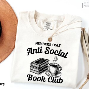 a white t-shirt with a graphic design that reads "MEMBERS ONLY Anti Social Book Club" and includes a book and a cup of coffee.