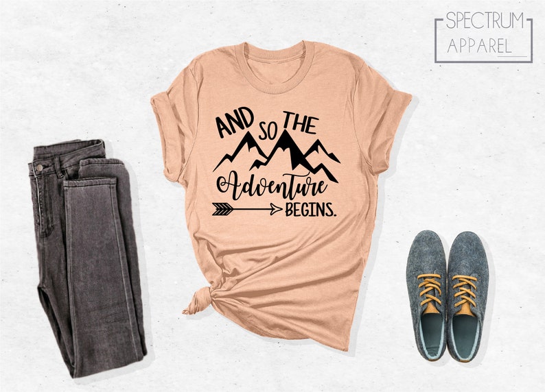 May include: A peach-colored t-shirt with a black graphic that says "And so the adventure begins." with a mountain range and arrow design.