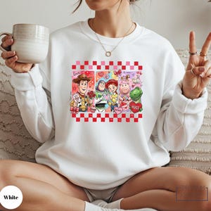 A woman wearing a white sweatshirt with a graphic of characters from Toy Story, including Woody, Buzz Lightyear, and other beloved characters, is sitting on a couch and holding a coffee mug.