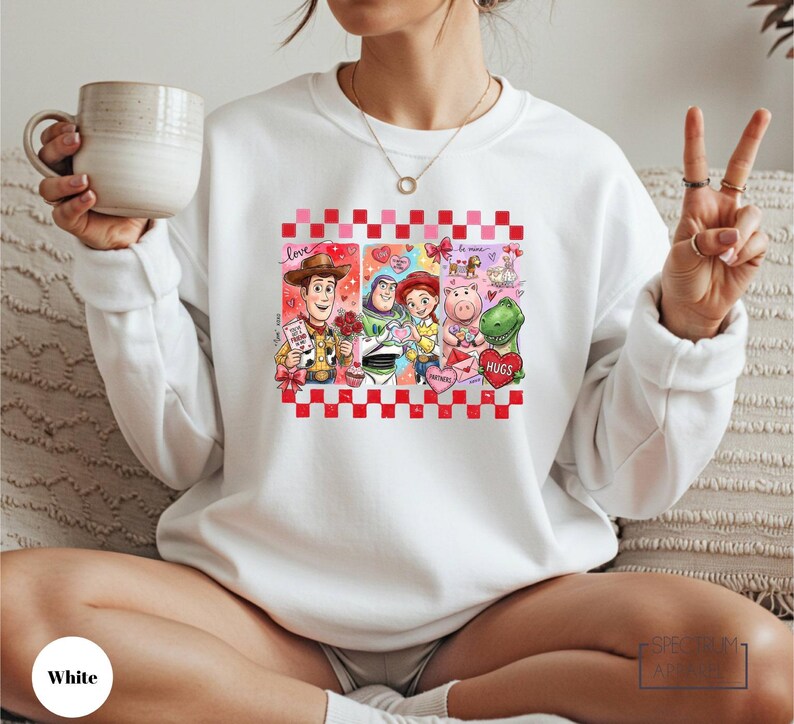 A woman wearing a white sweatshirt with a graphic of characters from Toy Story, including Woody, Buzz Lightyear, and other beloved characters, is sitting on a couch and holding a coffee mug.