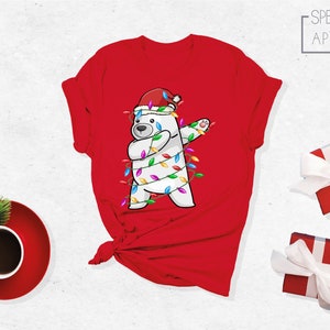 Op de afbeelding: Rood T-shirt met een cartoon ijsbeer met een kerstmuts en verpakt in kerstverlichting, in een dab-pose. Het shirt heeft de tekst "SPECTRUM APPAREL". Feestelijke kleding voor de feestdagen.