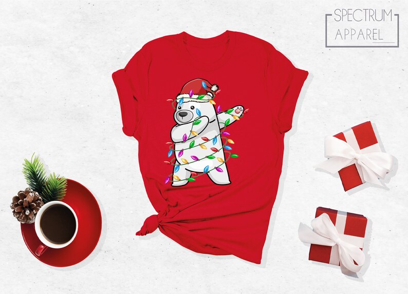 Op de afbeelding: Rood T-shirt met een cartoon ijsbeer met een kerstmuts en verpakt in kerstverlichting, in een dab-pose. Het shirt heeft de tekst "SPECTRUM APPAREL". Feestelijke kleding voor de feestdagen.