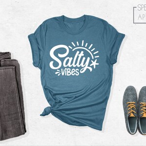 May include: A teal blue t-shirt with the text "Salty Vibes" in white lettering. The shirt is folded and tied in a knot. A pair of dark gray jeans and two gray shoes with orange laces are also pictured.