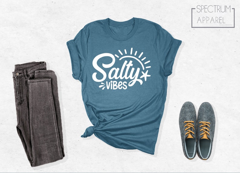 May include: A teal blue t-shirt with the text "Salty Vibes" in white lettering. The shirt is folded and tied in a knot. A pair of dark gray jeans and two gray shoes with orange laces are also pictured.