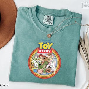 a light green t-shirt with a Disney Pixar logo, specifically Toy Story, printed on it. The t-shirt is laid out on a white surface, along with a brown hat and some dried leaves.