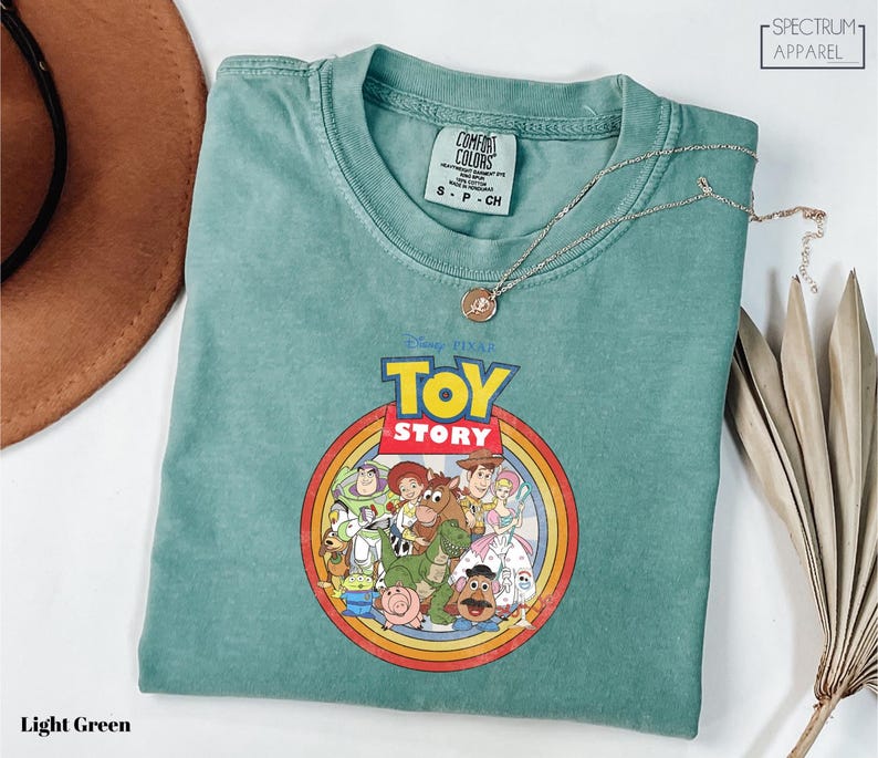 a light green t-shirt with a Disney Pixar logo, specifically Toy Story, printed on it. The t-shirt is laid out on a white surface, along with a brown hat and some dried leaves.