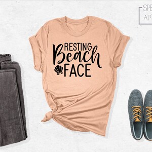 Może przedstawiać: Koszulka w kolorze brzoskwiniowym z napisem "Resting Beach Face" czarnymi literami i małym grafiką muszelki. Koszulka jest zawiązana na dole i jest prezentowana z parą ciemnoszarych dżins&oacute;w i dwoma szarymi butami ze złotymi sznur&oacute;wkami.