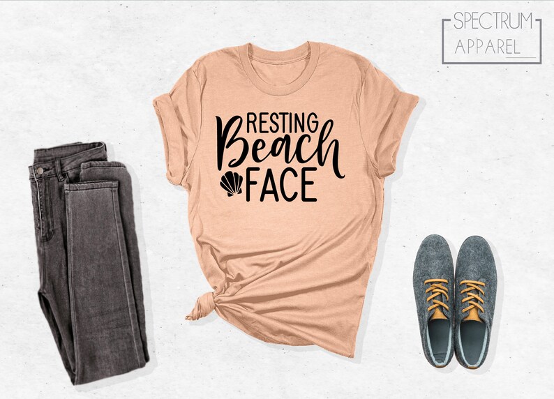 Może przedstawiać: Koszulka w kolorze brzoskwiniowym z napisem "Resting Beach Face" czarnymi literami i małym grafiką muszelki. Koszulka jest zawiązana na dole i jest prezentowana z parą ciemnoszarych dżins&oacute;w i dwoma szarymi butami ze złotymi sznur&oacute;wkami.