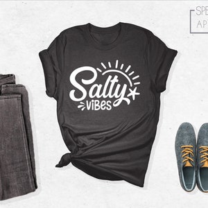 May include: A dark gray t-shirt with the text "Salty Vibes" in white lettering. The text is stylized with a sun and a starfish. The shirt is laid out on a white surface with a pair of dark gray jeans and a pair of gray shoes with orange laces.