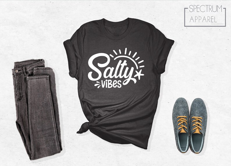 May include: A dark gray t-shirt with the text "Salty Vibes" in white lettering. The text is stylized with a sun and a starfish. The shirt is laid out on a white surface with a pair of dark gray jeans and a pair of gray shoes with orange laces.