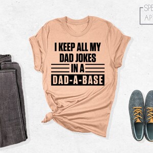May include: A peach-colored t-shirt with black text that reads "I keep all my dad jokes in a dad-a-base".