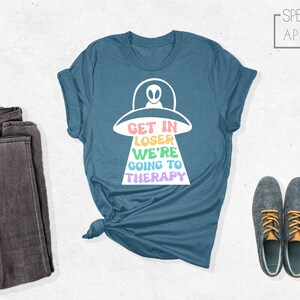 K&ouml;nnte beinhalten: Ein blaues T-Shirt mit einem wei&szlig;en Grafikdruck eines UFOs und dem Text "Get in Loser We're Going to Therapy" in Regenbogenfarben.