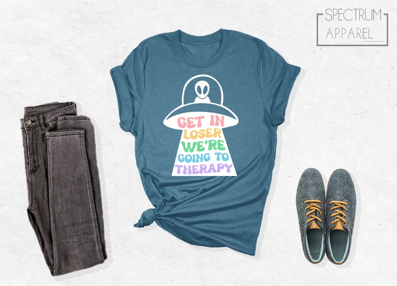 K&ouml;nnte beinhalten: Ein blaues T-Shirt mit einem wei&szlig;en Grafikdruck eines UFOs und dem Text "Get in Loser We're Going to Therapy" in Regenbogenfarben.