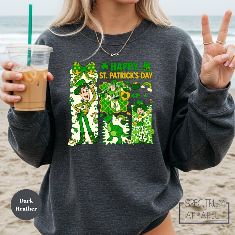 A woman wearing a gray sweatshirt with a graphic design featuring cartoon characters and text related to St. Patrick's Day. She is holding a drink in her hand and making a peace sign with her fingers.