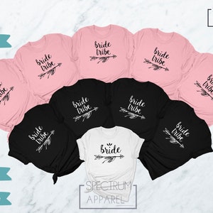 May include: Eight t-shirts in pink, black, and white with the text "bride tribe" and an arrow design. The shirts are arranged in a circle on a white surface.