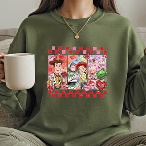A woman wearing a green sweatshirt with a graphic of characters from the Toy Story movie, including Woody, Buzz Lightyear, and Piggy, holding a white mug.