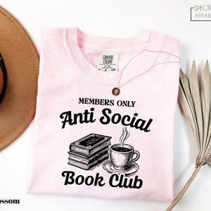 a pink t-shirt with a graphic design that says "MEMBERS ONLY Anti Social Book Club" and includes a book and a cup of coffee. The t-shirt is laid out on a white surface, with a brown hat and a plant visible in the background.